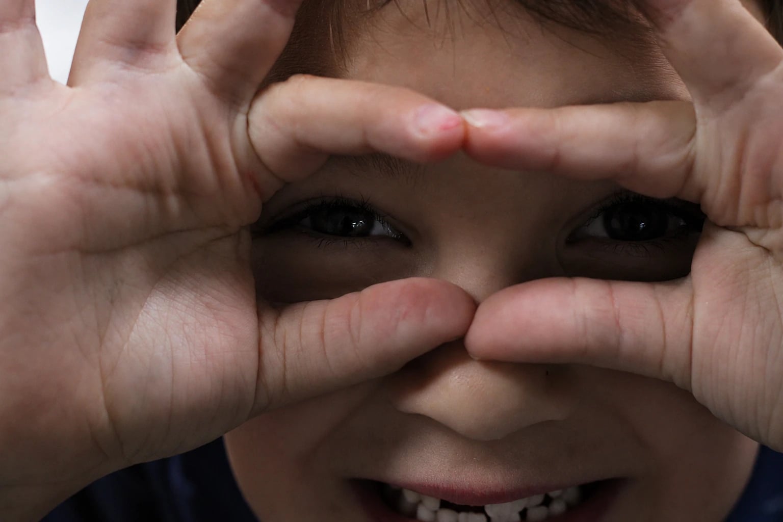 A boy grins through his fingers, held up like binoculars around his eyes.