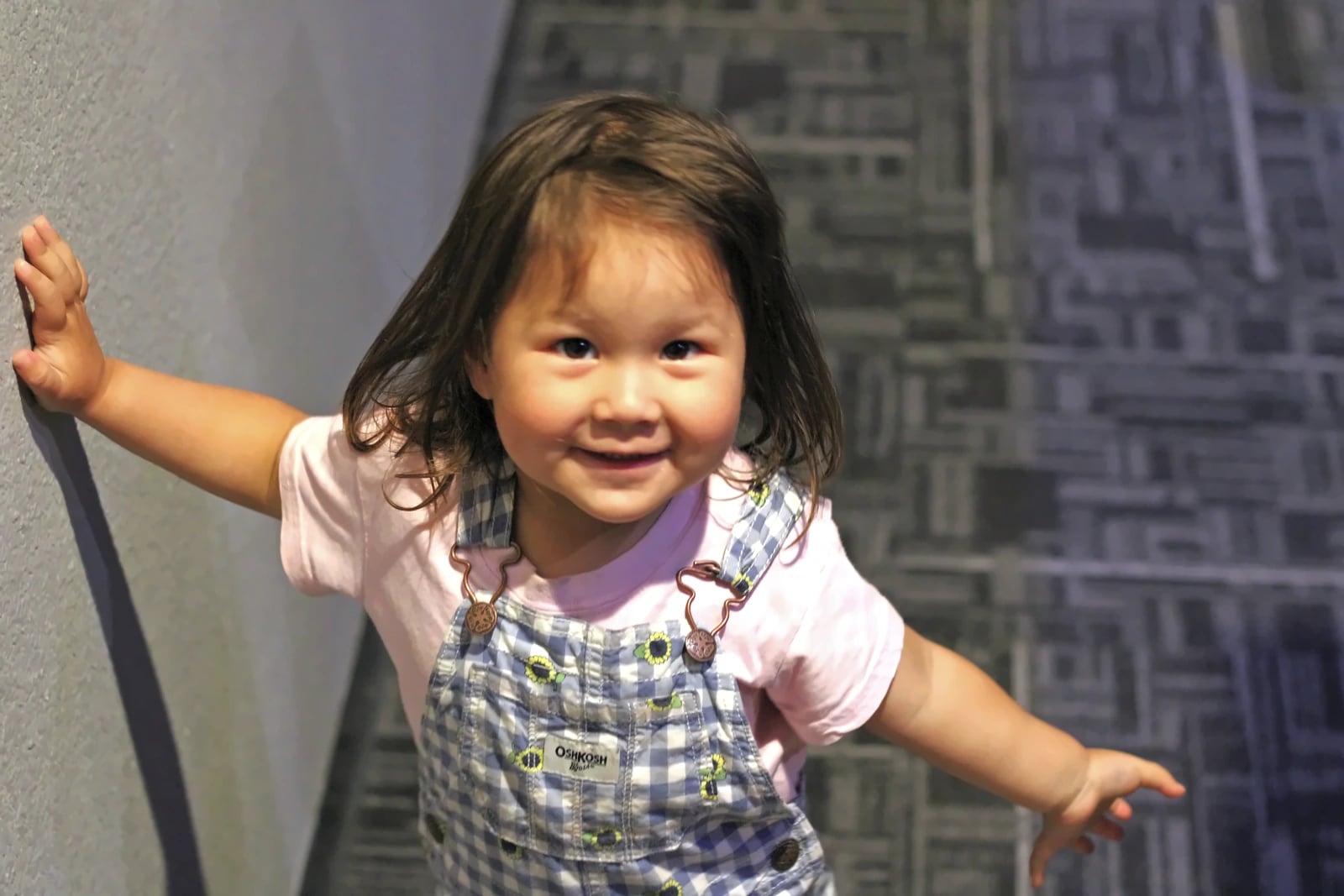 A toddler in overalls smiles in the church hallway.